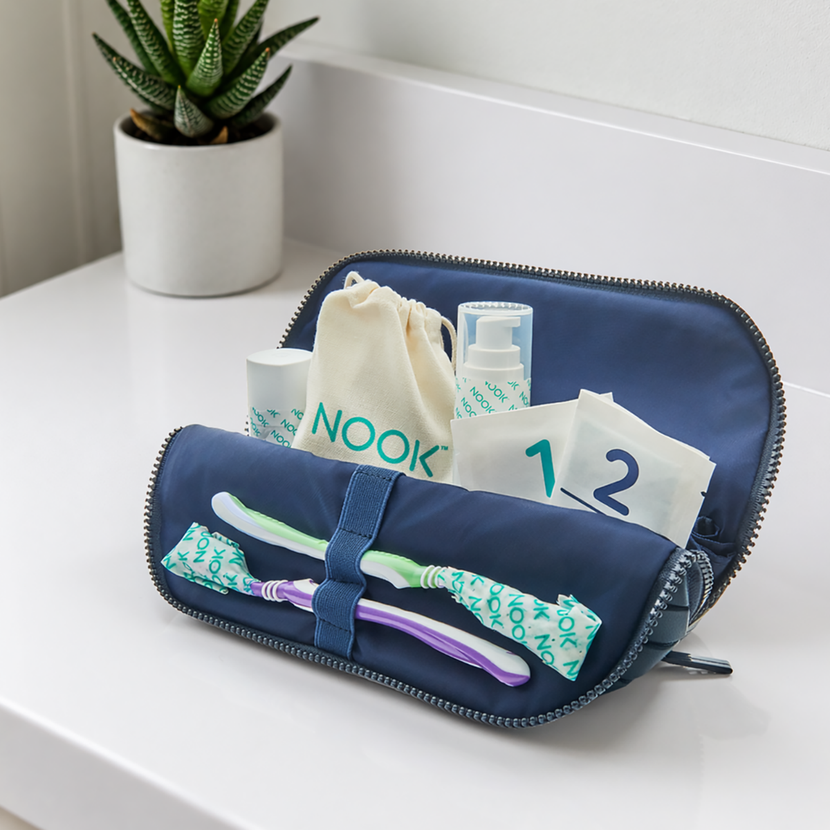 NOOK travel toiletry case open showing individually wrapped toothbrushes, a NOOK drawstring pouch, and travel bottles labeled 1 and 2