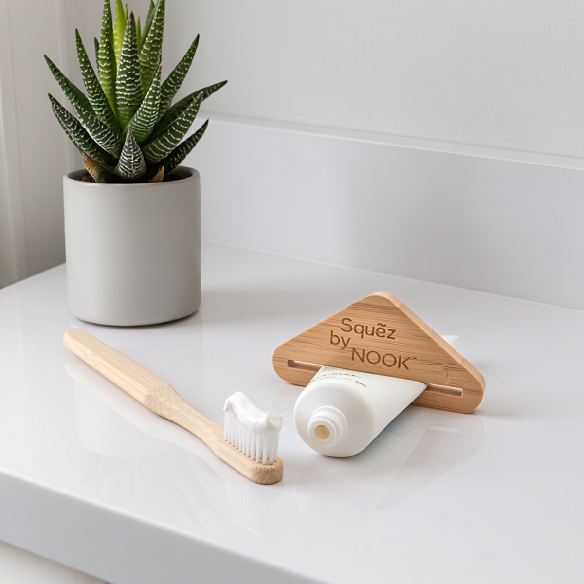 Squẽz by NOOK bamboo toothpaste squeezer, toothbrush, and toothpaste tube on a white bathroom counter with a small aloe plant