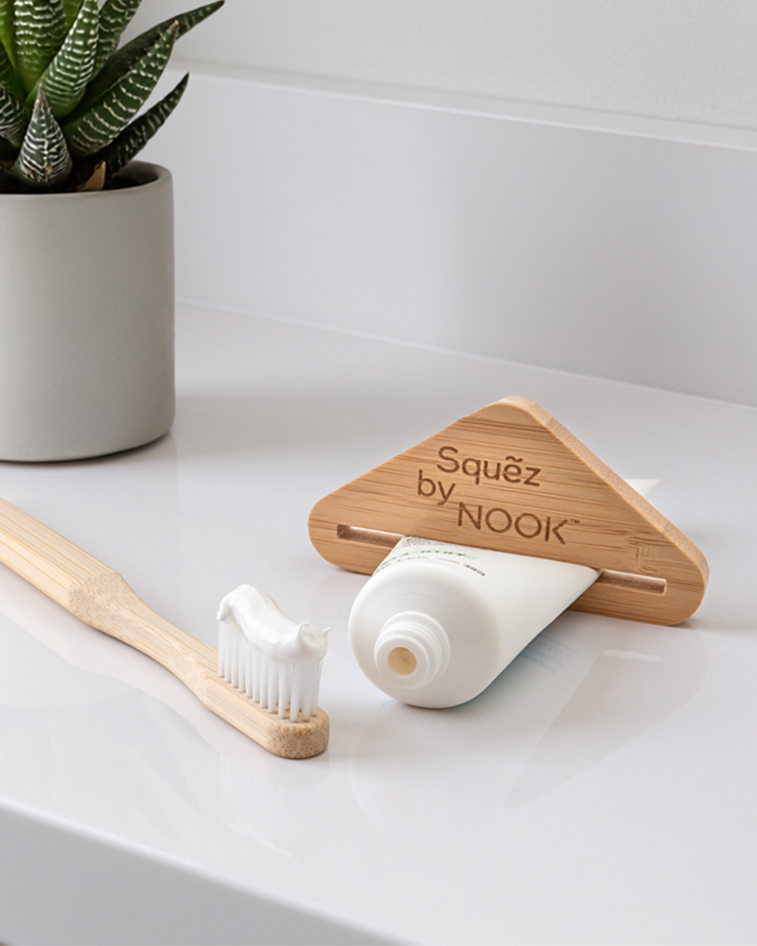 Squẽz by NOOK bamboo toothpaste squeezer with toothbrush and toothpaste on a white bathroom counter, vertical crop